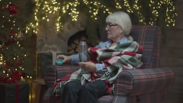 Старшая женщина в одеяле сидит в кресле у елки и пьет воду из бутылки в замедленной съемке . — стоковое видео