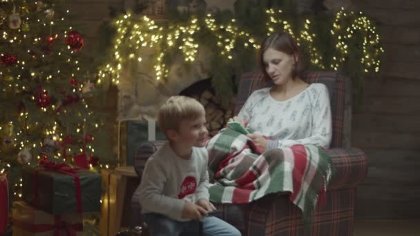 Ung kvinna i pyjamas i fåtölj surfa online på smartphone med blond son spelar på leksaksbil framför i juldekorationer i slow motion. — Stockvideo