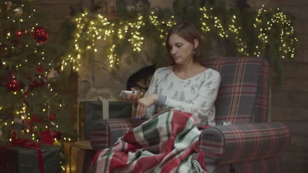 Jeune femme en pyjama dans un fauteuil en décoration de Noël frottant crème pour les mains. Brunette assise sur un canapé confortable à la veille de Noël — Video