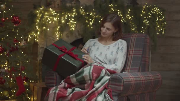 Mladá fena v pyžamu v křesle psaní jméno s perem na vánoční dárek s vánočním stromečkem a rozmazané světla na krbu vzadu ve zpomaleném filmu. — Stock video