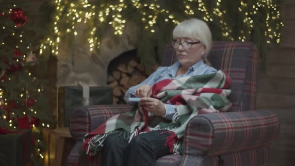 Seniorin in Decke sitzt im Sessel am Weihnachtsbaum und nimmt Tabletten mit Wasser in Zeitlupe. — Stockvideo