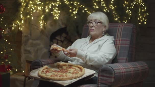 Senioren blonde vrouw die pizza eet op de fauteuil in kerstversiering in slow motion. Grootmoeder geniet van pizza bij open haard. — Stockvideo