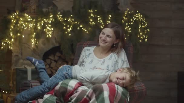 Молодая брюнетка 30-х годов, играющая с блондинистым сыном, сидящим в кресле в рождественском украшении. Счастливая женщина и мальчик смеются в замедленной съемке . — стоковое видео