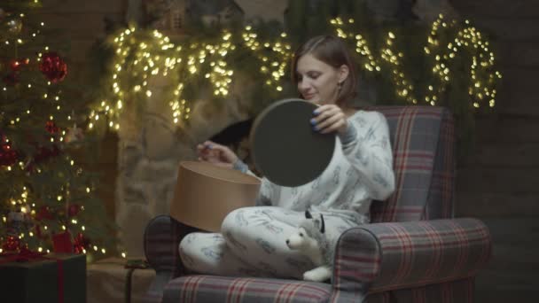 Jonge vrouw in pyjama 's in fauteuil die zacht puppy speelgoed maakt als kerstcadeau. Jonge vrouw zet speelgoed in huidige doos in Nieuwjaar decoratie in slow motion. — Stockvideo