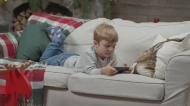 Bebek çocuk Noel süslemesinde şömineli akıllı telefon izliyor ve yılbaşı ağacını aydınlatıyor. Çocuk koltukta elinde aletle ağır çekimde Steadicam tarafından. 