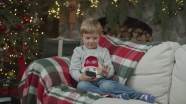 Bebek çocuk Noel süslemesinde şömineli akıllı telefon izliyor ve yılbaşı ağacını aydınlatıyor. Çocuk koltukta elinde aletle ağır çekimde Steadicam tarafından. 