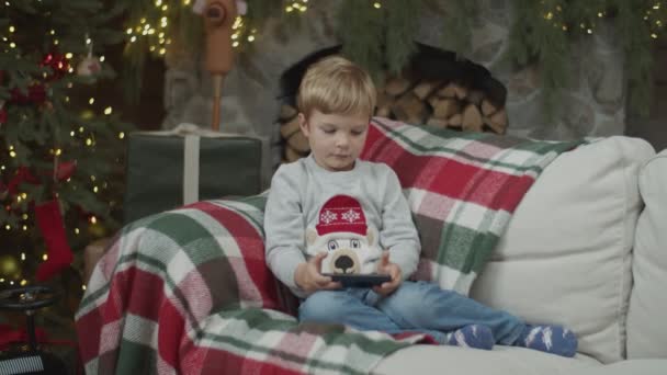 Niño viendo teléfono inteligente en la decoración de Navidad con chimenea y árbol de Navidad iluminado. Niño en sofá con gadget en cámara lenta por steadicam . — Vídeo de stock