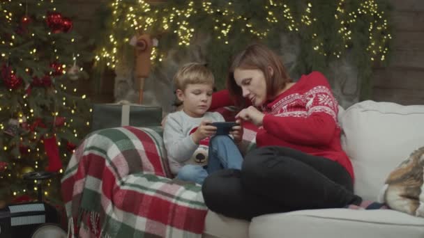 Morena mãe com filho loiro olhando para smartphone sentado no sofá na véspera de Natal. Família rindo perto da lareira em câmera lenta por steadicam . — Vídeo de Stock