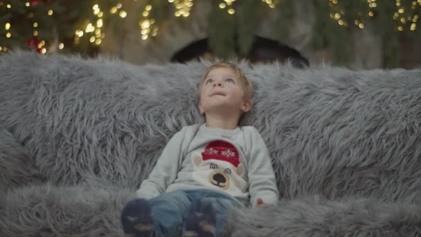 A kisfiú ül a szürke kanapén karácsonyi díszítésben lassított felvételen. A gyerek felemeli a kezét, és nem tudja.. — Stock videók