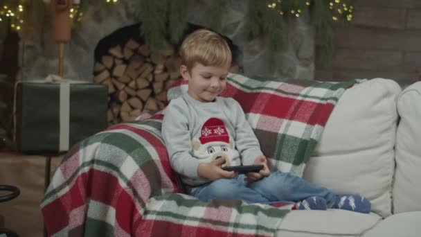 Baby pojke tittar på smartphone i juldekoration med öppen spis och tände julgran. Grabben på soffan med gadget i slow motion med steadicam. — Stockvideo