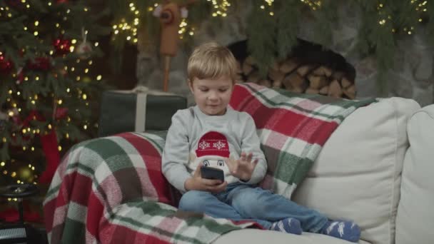 Bambino che guarda smartphone nella decorazione natalizia con camino e albero di Natale illuminato. Bambino sul divano con gadget al rallentatore di steadicam . — Video Stock