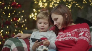 Gülümseyen esmer anne ve sarışın oğlun Noel arifesinde koltukta oturup akıllı telefona bakması. Aile şöminenin yanında gülüyor ağır çekimde. 