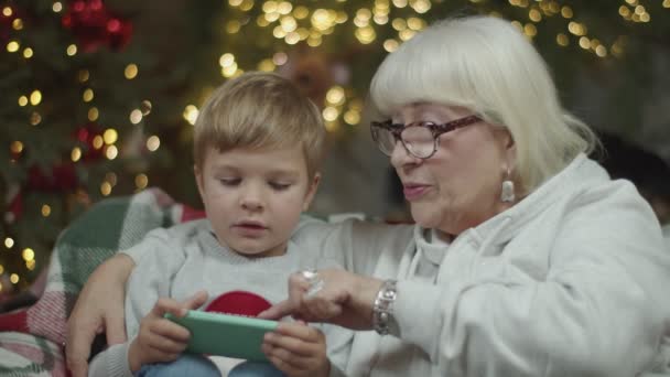 Primer plano de la abuela rubia en gafas con nieto mirando el teléfono inteligente sentado en el sofá cerca de la chimenea en las decoraciones de Navidad en cámara lenta . — Vídeos de Stock