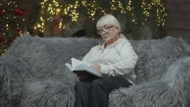 Seniorin mit Brille liest Papierbuch und sitzt an Heiligabend in Zeitlupe auf Sofa. — Stockvideo