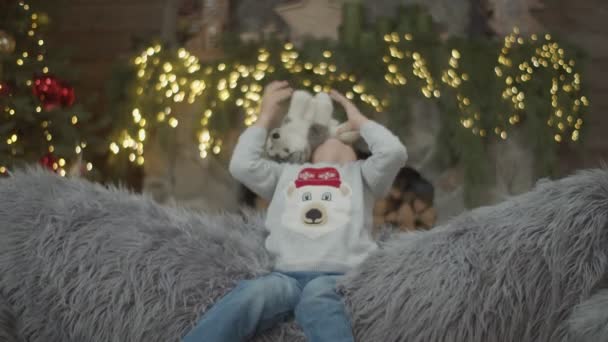 Der kleine Junge spielt in Zeitlupe mit Plüschtier auf dem grauen Sofa in Weihnachtsdekoration. Kind wirft und fängt Husky-Spielzeug. — Stockvideo