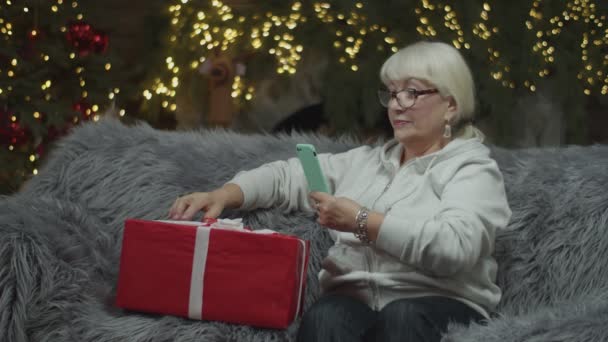 Senior mujeres rubias haciendo foto por teléfono de regalo de Navidad sentado en el sofá con luces borrosas en la chimenea detrás. Envoltura de regalo — Vídeos de Stock