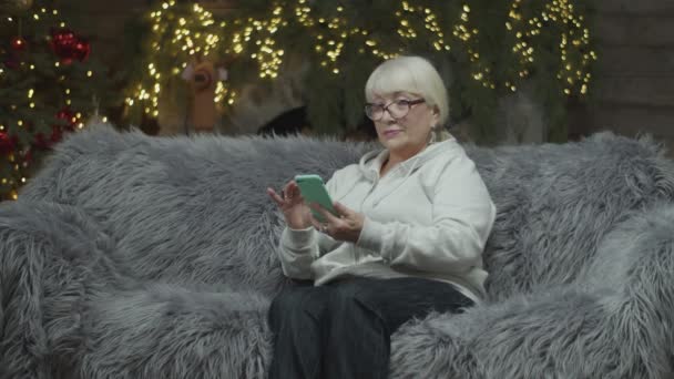 Seniorin surft an Heiligabend im Smartphone online und sitzt in Zeitlupe auf Sofa. — Stockvideo