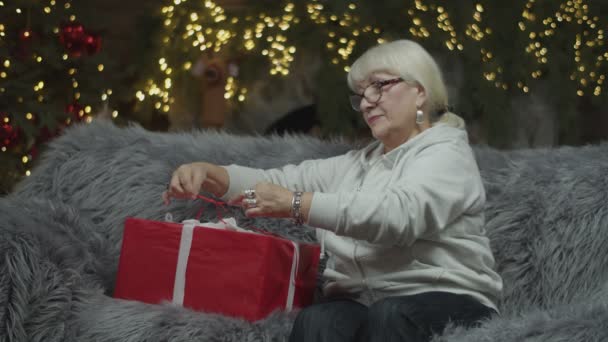 Senior mujeres rubias haciendo un lazo de rayas blancas para regalo de Navidad sentado en el sofá con luces borrosas en la chimenea detrás. Envoltura de regalo — Vídeo de stock