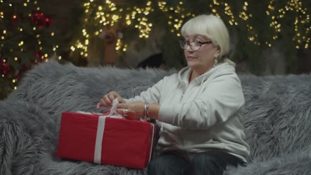 Senior mujeres rubias haciendo un lazo de rayas blancas para regalo de Navidad sentado en el sofá con luces borrosas en la chimenea detrás. Envoltura de regalo — Vídeo de stock