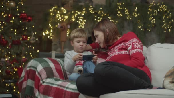 金髪の息子を持つブルネットの母親は、クリスマス・イブにソファに座っているスマートフォンを見ています。スローモーションで暖炉の近くで笑う家族. — ストック動画