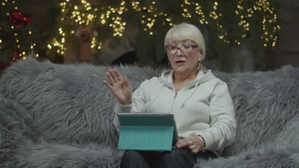 Senior rubia mujer hablando en línea por tableta con un amigo en Nochebuena sentado en el sofá en cámara lenta . — Vídeo de stock