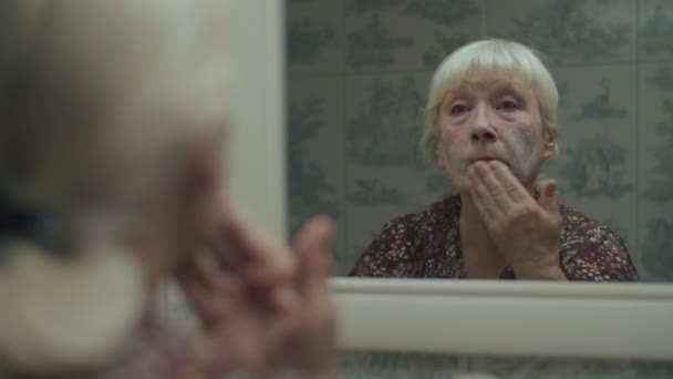 Senior fêmea loira aplicando creme hidratante e olhando no espelho de reflexão em câmera lenta. Mulher idosa aplicando creme com as mãos no rosto . — Vídeo de Stock
