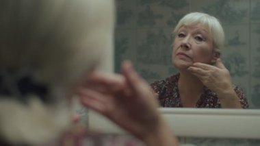 Yaşlı sarışın kadın yavaş çekimde ayna yansımasına makyaj yapıyor. Yaşlı bir kadın elleriyle yüzüne temel atıyor.. 
