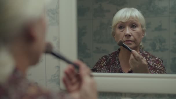Die ältere blonde Frau schminkt sich in Spiegelreflexion in Zeitlupe. Seniorin sprüht Puder mit Frechheit ins Gesicht. — Stockvideo
