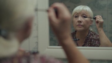 Yaşlı sarışın kadın yavaş çekimde ayna yansımasına makyaj yapıyor. Kirpik için rimel süren yaşlı bir kadın.