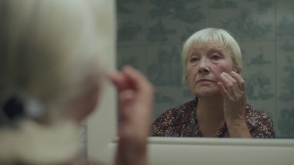 Idősebb szőke nő, aki hidratáló krémet használ, és lassított felvételen tükörképpel néz a tükörbe. Idős nő krémet használ kezekkel az arcán. — Stock videók
