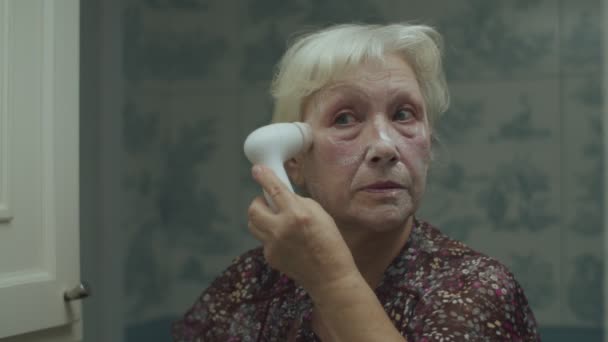 Mulher loira sênior fazendo massagem facial com máquina em espelho de reflexão em câmera lenta. Mulher idosa usando máquina de mão para massagem facial . — Vídeo de Stock