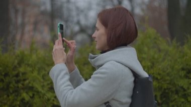 Esmer kadın, sonbahar parkında akıllı telefonuyla şehir görüşünün fotoğrafını çekiyor. Sırt çantalı, üniversitenin fotoğrafını çeken gri ceketli kadın turistin yan görüntüsü.. 