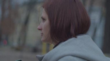 Genç esmer anne, sonbahar parkında oyun parkının yanında oturan akıllı telefonuna bakıyor. Parkta telefonla yürüyen gri ceketli kadınlar.. 