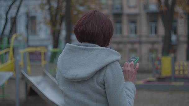 Mladá brunetka matka se dívá na svůj smartphone sedí u hřiště v podzimním parku. Ženy v šedém kabátě s telefonem procházky v parku. — Stock video