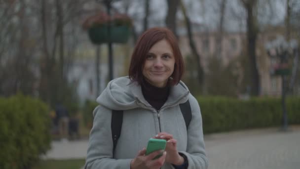 갈색 머리 여자가 가을 공원에서 스마트폰으로 도시 풍경을 찍고 있습니다. 앞을 보고 있죠. 대학 사진을 찍고 있는 가방을 든 회색 코트를 입고 있는 여자 관광객. — 비디오