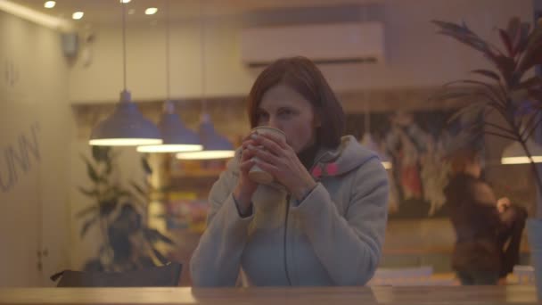 Mujeres jóvenes en abrigo bebiendo café en la cafetería, ventana disparada en cámara lenta. Mujer sentada junto a la ventana en coffeeshop con taza de bebida . — Vídeo de stock