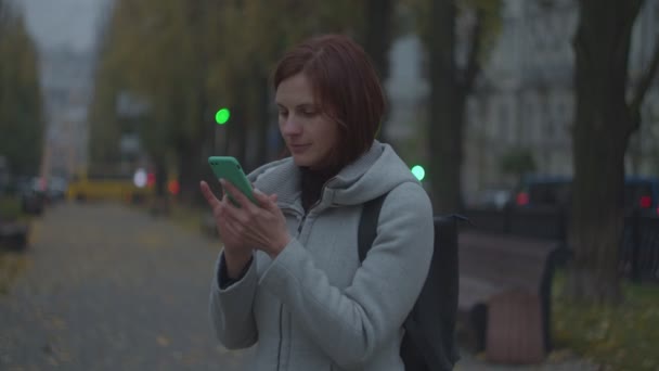 Brunette woman tourist in gray coat surfing online in smartphone in fall evening park with blurred city behind in slow motion. — Stock Video