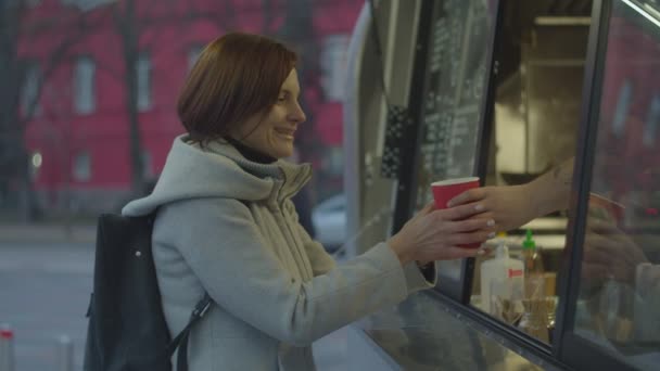 Young brunette female with backpack buying hot coffee drink at street food truck. Woman feeling happy to get hot tea in cold hands. — ストック動画