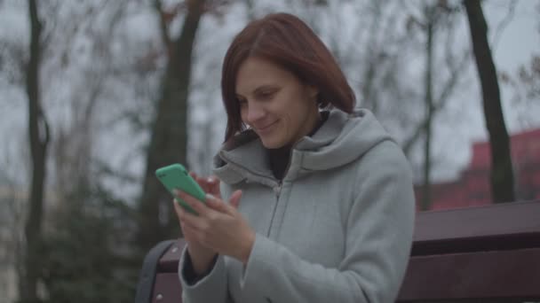 Bruna femmina adulta con smartphone seduta su panchina nel parco autunnale. Donna sorridente durante la navigazione online sul telefono . — Video Stock