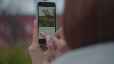 Esmer kadın, sonbahar parkında akıllı telefonuyla şehir görüşünün fotoğrafını çekiyor. Gri ceketli, sırt çantalı, üniversitenin fotoğrafını çeken bir kadın turist.. 