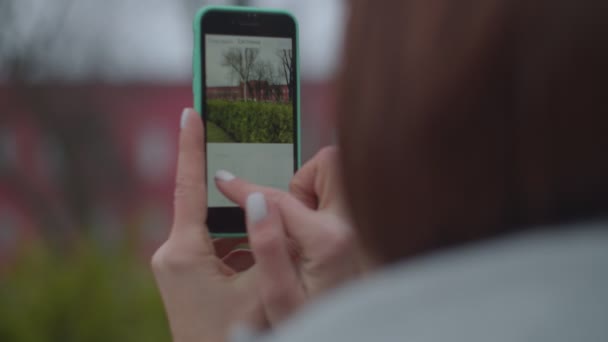 Mujer morena tomando fotos de la vista de la ciudad en su teléfono inteligente en el parque de otoño. Turista femenina en abrigo gris con mochila haciendo foto de la universidad . — Vídeo de stock