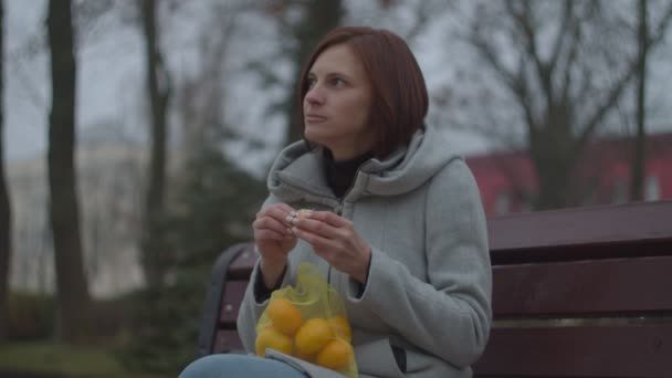 Ung brunett kvinna peeling mandariner på bänken i höstparken. Kvinna med miljövänlig väska med mandariner leende medan du äter mandariner. — Stockvideo
