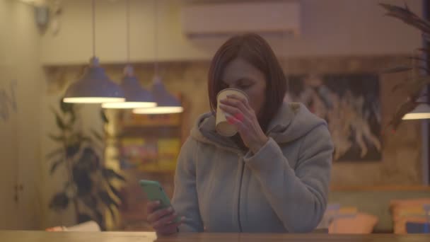 Las mujeres jóvenes tomando café en la cafetería y charlando en línea en el teléfono inteligente, ventana de disparo en cámara lenta. Copa en manos femeninas . — Vídeo de stock