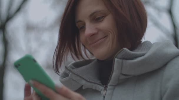 Крупный план брюнетки взрослой женщины со смартфоном, улыбающимся в осеннем парке в замедленной съемке . — стоковое видео