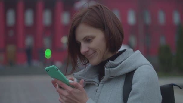Bruna donna turista in cappotto grigio navigare online in smartphone in autunno parco con università dietro al rallentatore . — Video Stock