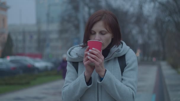 Mladé ženy pijí horkou kávu nebo čaj v červeném papírovém kelímku na větrné ulici. Žena cítí chlad a drží horký šálek kávy v rukou. — Stock video