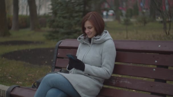 Brunette female adult with smartphone sitting on bench in fall park. Woman smiling while surfing online on phone. — ストック動画