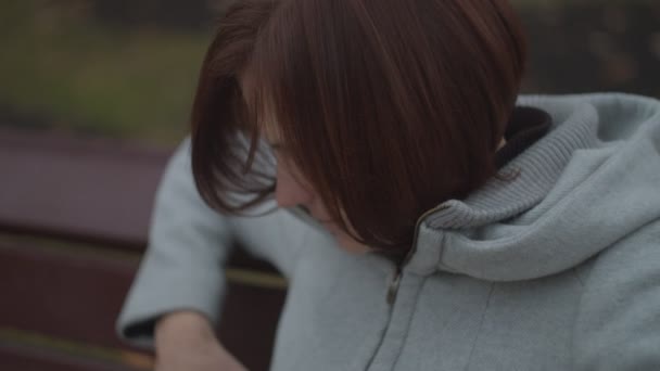 스마트폰을 들고 가을 공원에 벤치에 앉아 있는 갈색 머리 여자 어른. 온라인 서핑을 하며 웃고 있는 여성. — 비디오