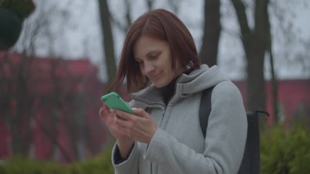 Brunette kvinde turist i grå frakke surfing online i smartphone i efteråret park med universitet på bagsiden i slowmotion . – Stock-video