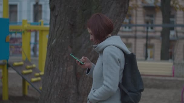 Giovane madre bruna guardando il suo smartphone seduto vicino al parco giochi nel parco autunnale. Donne in cappotto grigio con telefono che cammina nel parco . — Video Stock